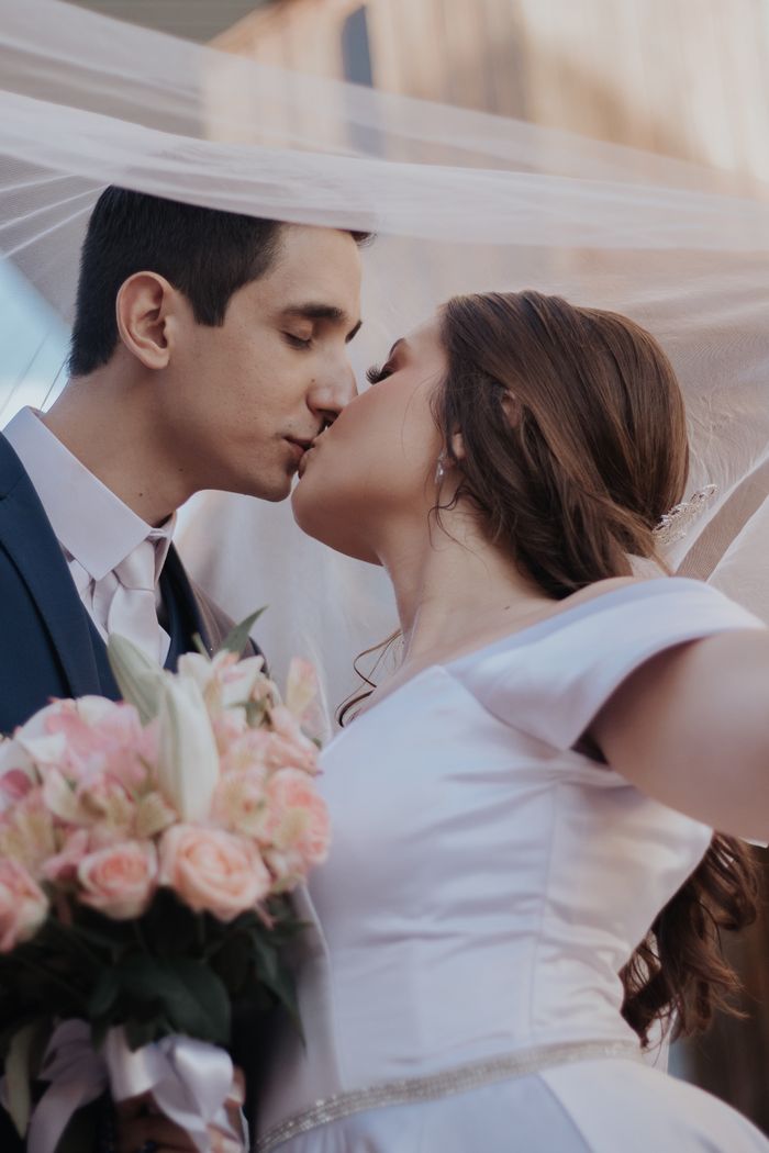 Bride and groom share a tender kiss under a veil, holding a bouquet of flowers.